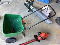 Photo showing Scotts seed spreader and electric hedge trimmer on garage floor, spreader has green hopper and black wheels, trimmer red and black body