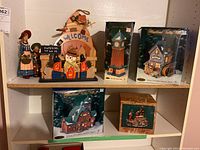 Photo of multiple holiday figurines and boxed Heartland Valley Village lighted houses and musical figurines displayed on shelving for lot identification.