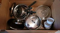 Photo of cardboard box containing Pyrex dishes, ceramic ramekins, stainless steel saucepans and pots, frying pan, and lids