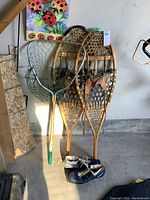 Photo shows a pair of traditional wooden snowshoes with rawhide webbing, a 39-inch fishing net with a green handle, and a pair of blue and white cross country ski shoes on the floor under a stairway.
