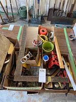 Overview of tools and hardware including large metal squares, rulers, clips, rolls of caution tape, wire brushes, and assorted small hardware organized in boxes on a wooden table.