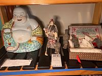 Shelf display of multiple Asian figurines including Laughing Buddha, standing figure, and boxed tomb figurines.