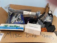 Photo of a cardboard box labeled 'Misc Bike Parts' containing boxed Wellgo pedals, Easton bike parts, Dura-Ace part box, CycleOps accessory, reflectors and miscellaneous bicycle components.