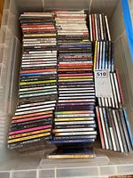 Top view of plastic bin filled with stacked rows of jewel case CDs. Variety of artists and genres visible, all tightly packed.