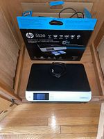HP Envy 5530 printer placed on wooden floor next to its original box. Power cable coiled on the printer.
