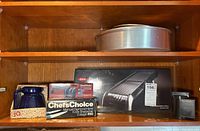 Kitchen shelf displaying all items together showing boxed mandolin slicer, knife sharpener, blue ceramic grinder, and bakeware pans.