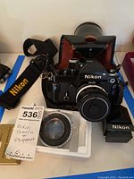 Photo showing Nikon manual film camera mounted with a Nikkor 50mm f/1.4 lens, black Nikon camera strap with yellow lettering, black leather Nikon camera case, type C focusing screen in white packaging, and handwritten label noting Nikon camera and equipment.