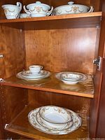 Noritake china pieces including sugar bowl, creamer, covered serving bowl on top shelf and stacked bowls and plates on middle and lower shelves in a wood cabinet.