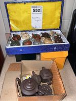 Blue fabric-covered box open to reveal eleven miniature teapots; cardboard box below with additional pots