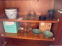 Shelf display showing the Tiffany glass bowl in its original box, two green decorative bowls with designs, ceramic planter, two brass pinecone decor pieces, and the small wooden stands.