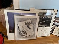 Overview of four stacked wall art pieces leaning against a wall, including a framed Escher print and Ansel Adams Gallery posters