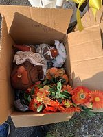 Open cardboard box containing two fabric turkeys and artificial autumn flowers in orange, yellow, and green colors.