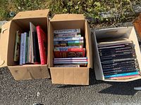 Three open boxes filled with books. Left box contains various cookbooks and reference books with colorful spines, the middle box prominently shows cookbooks with visible titles, right box contains mostly black-covered travel or reference books with white titles.