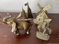 Four brass decorative items: windmill doorstop, sailboat, elephant, man on donkey on wooden surface