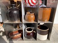 Ceramic crocks and vessels arranged in metal shelving cubby compartments, various sizes and colors including brown glazed jug and cream and brown crocks.