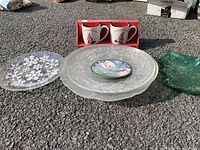 Display of four Christmas-themed glass platters and two red boxed ceramic mugs on outdoor ground.