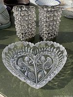 Two heart-shaped metal candle sticks with circular crystal-like embellishments and a clear glass heart-shaped trinket plate with embossed bird and leaf pattern