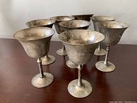 Group photo showing eight tarnished metal goblets arranged in two rows on a wooden surface, with visible signs of age and dirt needing cleaning.