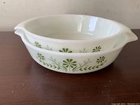 Single white milk glass casserole dish with green floral and leaf pattern around sides, round shape with two spouts on opposite sides on a wooden surface