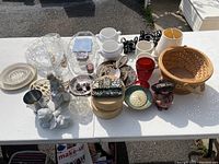 Wide view of various items on a white table: glass plates and bowls, ceramic boxes, wooden boxes, metal candle holders, figurines, woven basket, and decorative items.