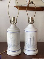 Pair of white ceramic lamps with brass harp frames, showing schooner sketches and the name Dancing Feather on the bases. Lamps have no bulbs or shades.