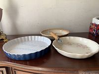 Three ceramic pie plates including one with blue text, floral pattern, and pumpkin pie theme, alongside a wooden handled pie edge cutter.