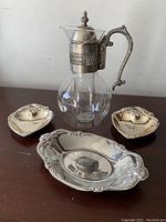 Full view of glass carafe with metal lid and handle, flanked by three silver metal dishes including a large oval tray and two small ornamental dishes.