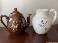 Brown ceramic teapot with decorative details next to white pitcher with purple flower design and twisted handle on a flat surface.