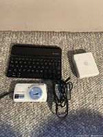 Photo showing the Nikon Coolpix camera, Logitech keyboard case, and Apple power adapter placed together on a carpeted floor.