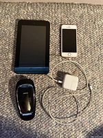 Photo showing a black tablet, a white iPhone, a white charger with cable, and a black Jabra Bluetooth headset on a fabric background.