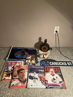 Photo shows vintage Vancouver Canucks game programs, Canucks flag, bear figurines, NHL lamp, street sign, and wall applique arranged on floor.