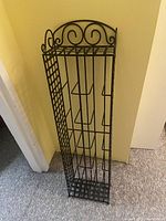 Full view of the black metal CD stand showing scroll design, vertical slots, and grid shelves, positioned against yellow walls on a carpeted floor.