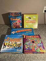 Photo showing five children's board games arranged on the floor against a beige wall: Pirate! The Octopus Sets to Sea, Whatzit? Junior, Pictionary Junior, Scrabble Junior, and Junior Serpent & Ladders.