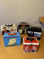 Front view of six metal lunch boxes showing Elvis Presley, AC/DC, Curious George, Coca-Cola, and U2 designs