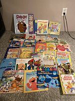 Photo showing full assortment of 26 colorful children's books spread on carpet. Titles visible include Scooby-Doo, Caillou, Arthur, Blue's Clues, and more.