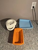 Photo showing white Cloer mini donut maker, orange rectangular silicone pan and blue square silicone pan on carpet, near outlet, side view.