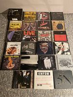 Top-down image showing 23 rock music CDs arranged in a grid, featuring covers by bands such as R.E.M., Ramones, Papa Roach, KMFDM, 5N, and L.A. Guns.