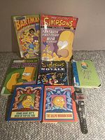 Full lot shows seven Simpsons-themed books and a Simpsons character wristwatch resting on carpet