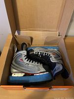 Pair of Bauer Vapor XVI hockey skates inside an orange box, showing worn condition, silver and black with visible signs of use and blue skate guards on the blades.