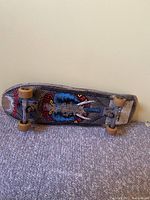Photo of entire vintage skateboard laying on carpet with graphic side facing up showing wear, wheels, and trucks.