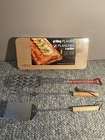 Photo of unopened package of 6 wooden grilling planks and three BBQ tools including a metal grilling fork, basket, and spatula with wooden handles.