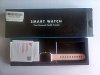 Photo of the smart watch and original box, black silicone strap, and included USB charger cable laid out on a white surface.