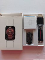 Photo showing the smart watch, USB charging cable, user manual, and original box packaging displaying the watch face.