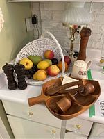 Photo showing white wicker basket filled with assorted faux fruit, wooden pepper mill, ceramic salt shaker, lamp with brass base and white glass shade, and wooden divided dish with utensils.