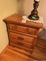 Front view of first wooden bedside table with brass handle drawers and brass lamp base on top.
