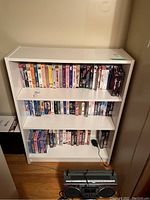 Photo showing front view of white bookcase filled with VHS tapes on three shelves, wooden floor background.