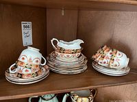 Photo showing the full china set including stacked saucers, dessert plates, sugar bowl and creamer with tea cups arranged, all featuring detailed orange and gold floral patterns