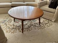 Round wooden coffee table with polished surface and turned legs on a carpet near upholstered seating.