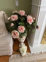 Full view of ornate vase filled with faux pink and white peony flowers beside a beige armchair and white fireplace.