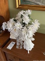Photo showing white faux flowers in rectangular white container with bisque and glass figurines on sides